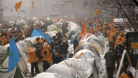Tents and protestors in orange jackets covered in snow, flags surround them